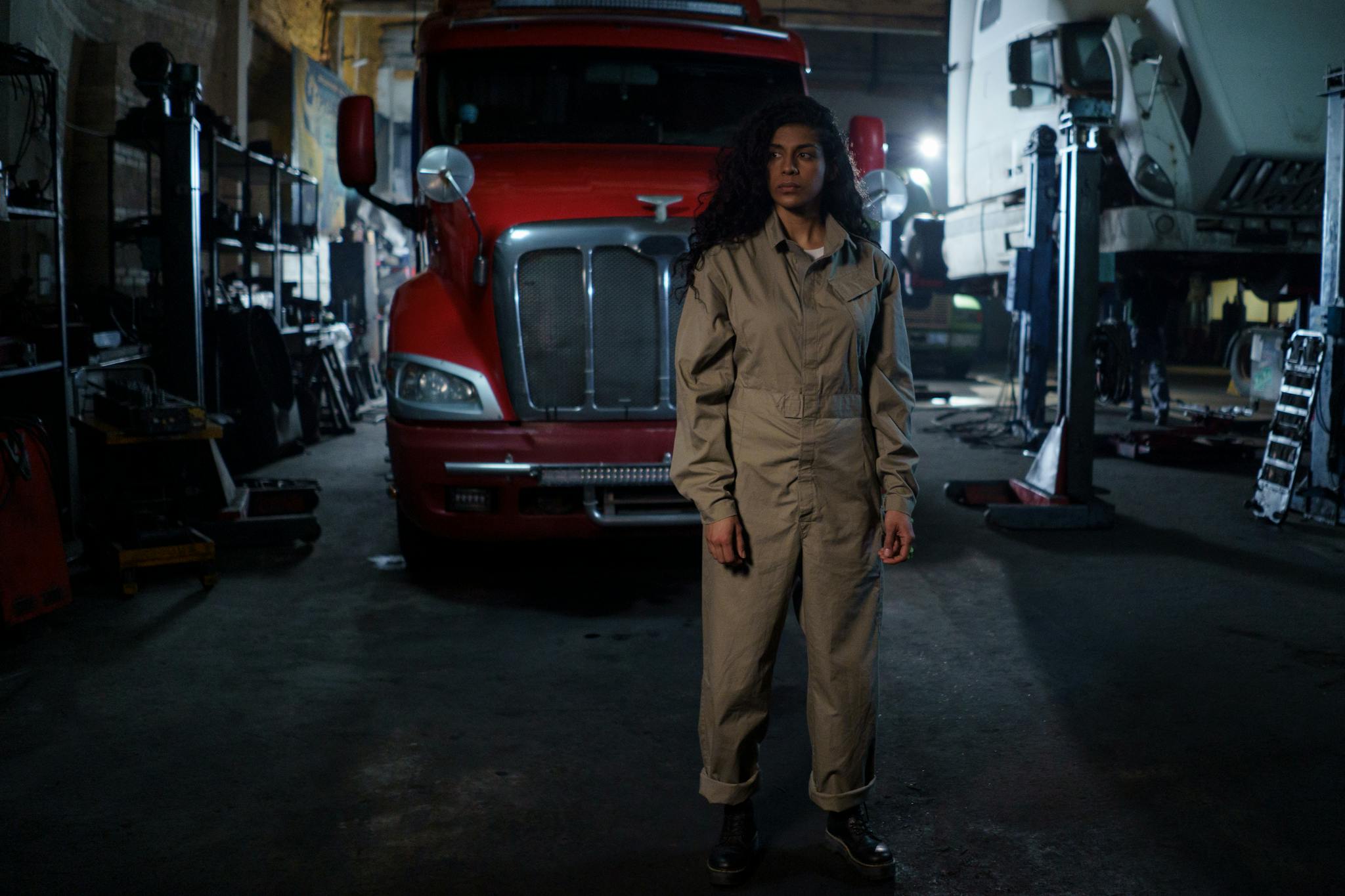 A woman in a uniform stands confidently in an automotive repair shop with trucks around.