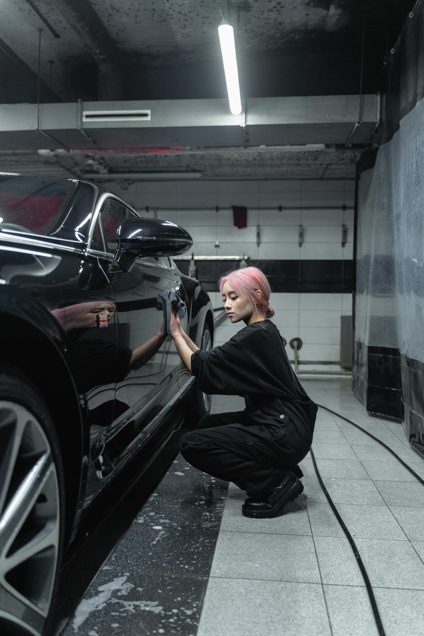 A woman with pink hair cleaning a black car in an indoor garage setting.